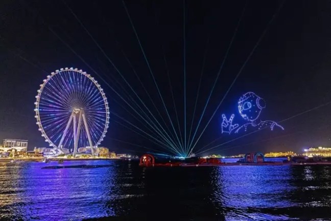 Ferris wheel and drone light show over water at night, with a digital character waving.