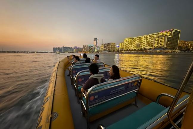 People on a boat cruising at sunset with city skyline and illuminated buildings in the background.