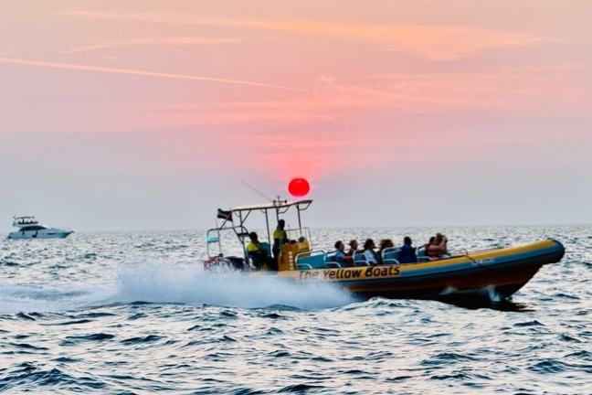 Speedboat with passengers on the sea at sunset with a distant yacht in view.