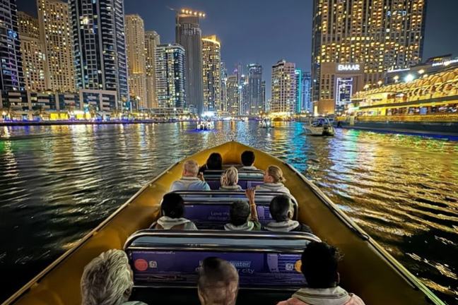 People on a boat at night in a city with illuminated skyscrapers reflecting on the water.